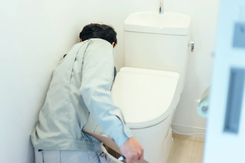 Person in work clothes inspecting or fixing a white toilet in a bathroom corner.