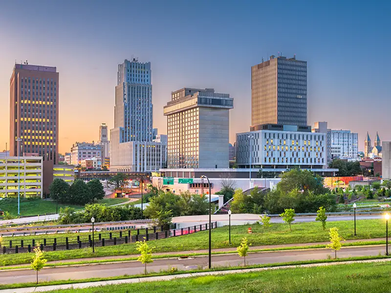City skyline with tall buildings at sunset behind a green park and roads.
