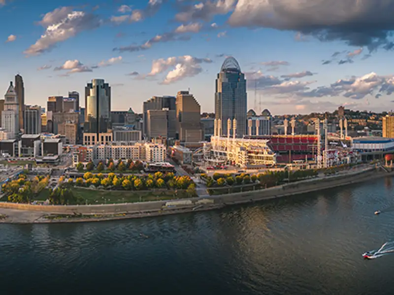 City skyline with a riverfront park and a large stadium under a partly cloudy sky.