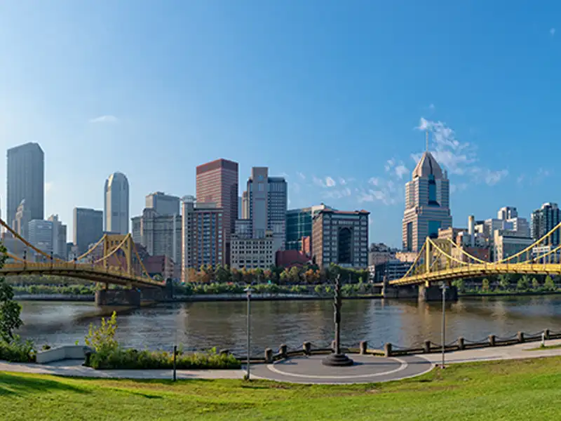City skyline with two yellow suspension bridges over a river and a statue in the foreground.