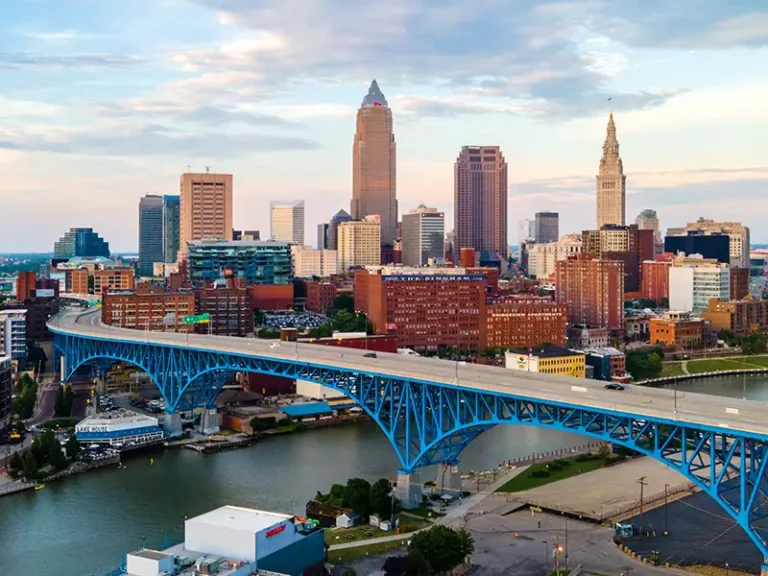 Blue steel bridge spanning a river with a city skyline in the background at dusk.