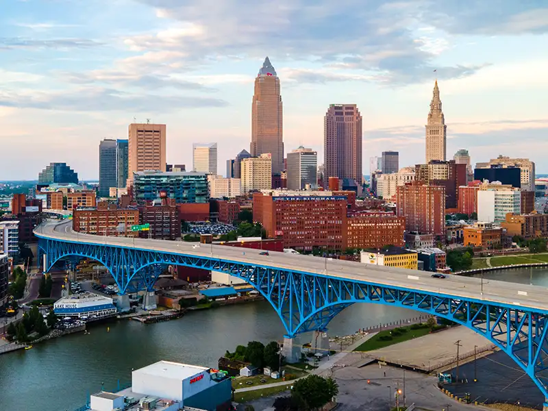 Blue steel bridge spanning a river with a city skyline in the background at dusk.