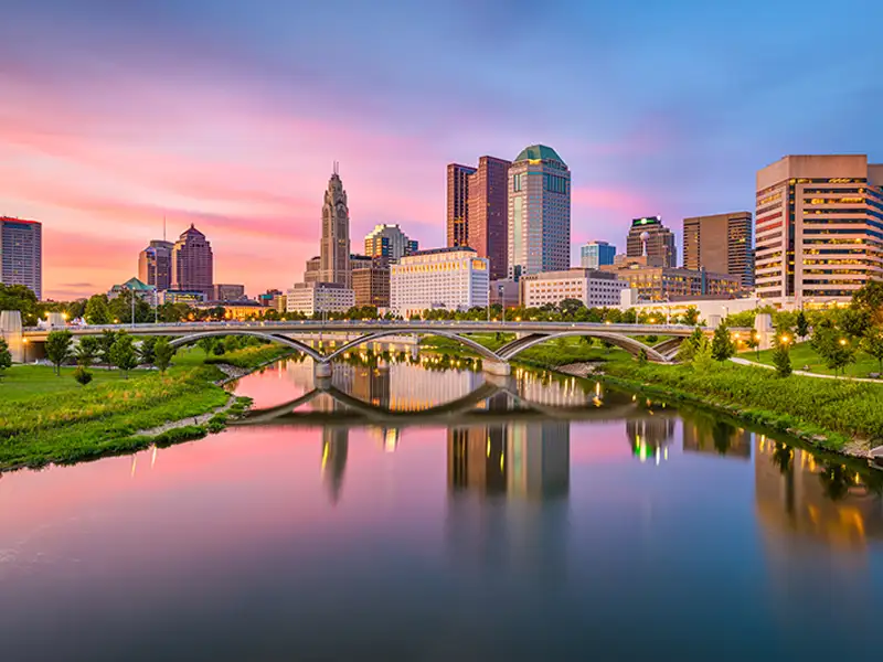 City skyline with a bridge over a calm river at sunset, reflecting colorful sky and buildings.