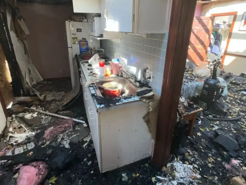 Kitchen and adjacent room severely damaged by fire with charred debris and soot covering surfaces and floor.