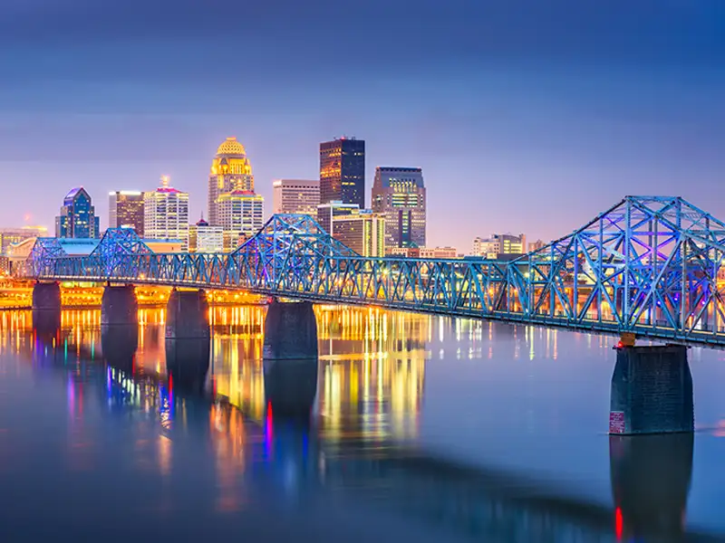 Blue-lit bridge over calm river with a city skyline illuminated at dusk in the background.