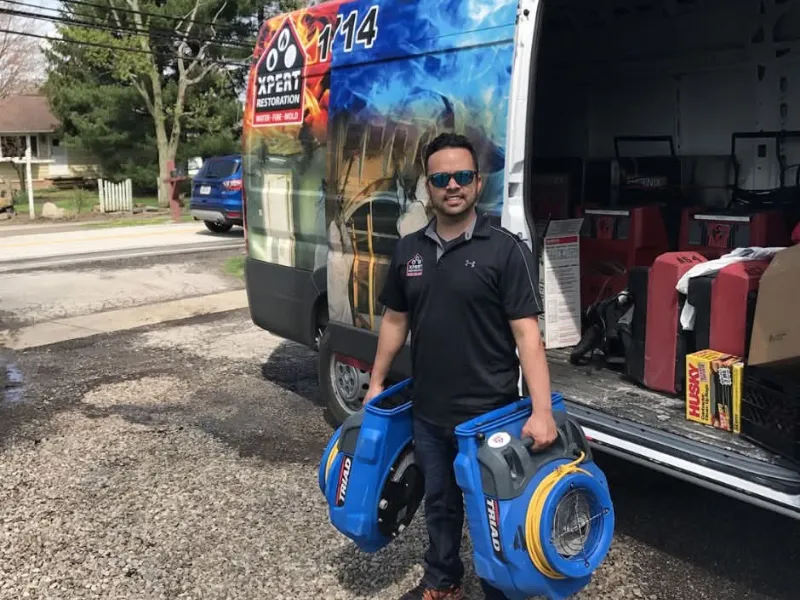 Man wearing sunglasses holding two blue industrial fans standing next to a restoration service van.