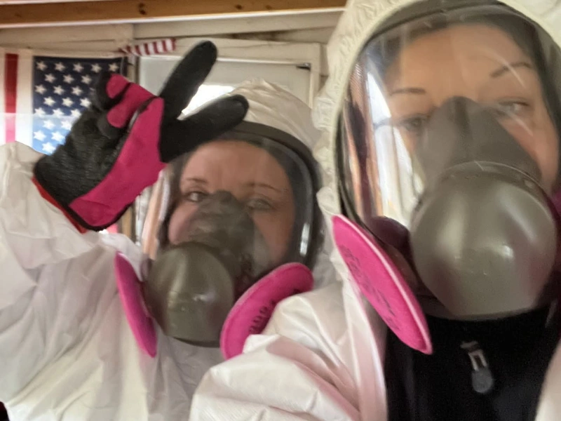 Two people wearing white hazmat suits and gas masks, one making a peace sign.