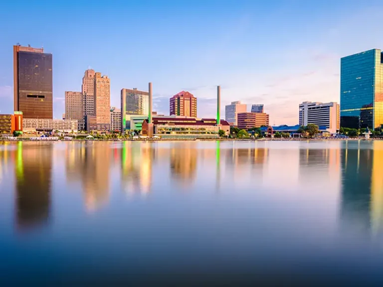 City skyline with buildings reflecting on calm water at sunset.